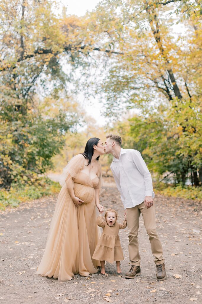 Abigail in cream maternity gown holding her baby bump outdoors in fall leaves with husband Owen and daughter Lainey in coordinating cream outfits.